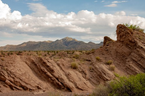 A rugged desert landscape with rocky formations and distant mountains under a partly cloudy sky.