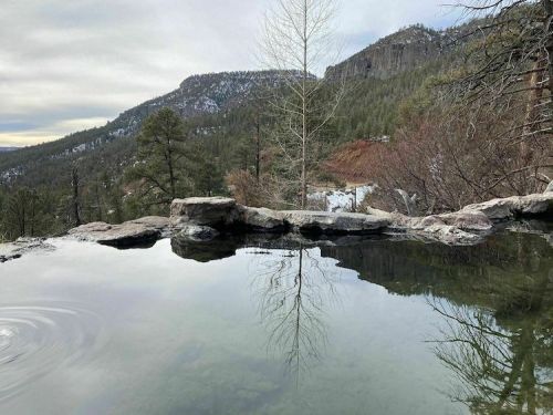 A serene hot spring surrounded by trees and mountains, reflecting the landscape in calm water.
