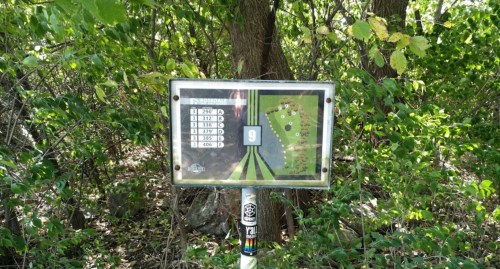A sign in a wooded area displaying a map and details for hole 9 of a disc golf course.