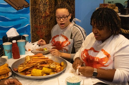 Two women wearing bibs enjoy a seafood feast with crab, corn, and drinks at a lively restaurant setting.