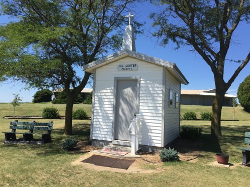 A small white chapel with a cross on top, surrounded by trees and benches, set against a clear blue sky.