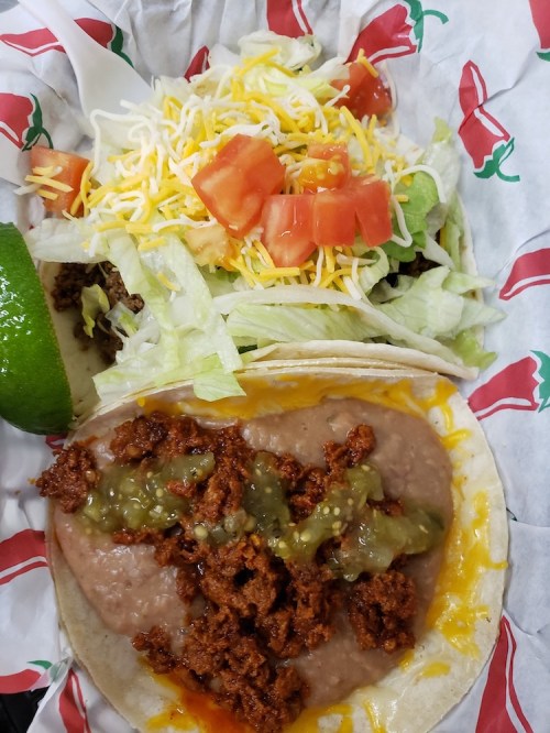 A close-up of two tacos with seasoned meat, refried beans, cheese, lettuce, tomatoes, and a lime wedge on a festive wrapper.