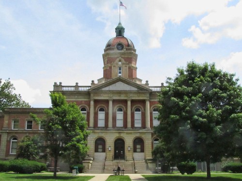 Historic building with a dome and clock tower, surrounded by trees and a grassy area under a blue sky.