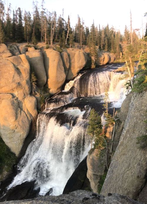 A scenic waterfall cascading over rocky cliffs, surrounded by lush trees and bathed in warm sunlight.