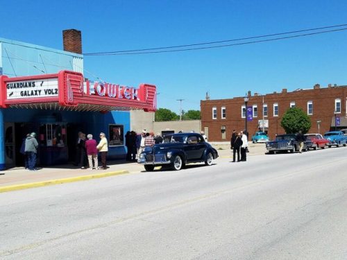 A vintage car parked outside a movie theater with people lined up for a film, under a clear blue sky.