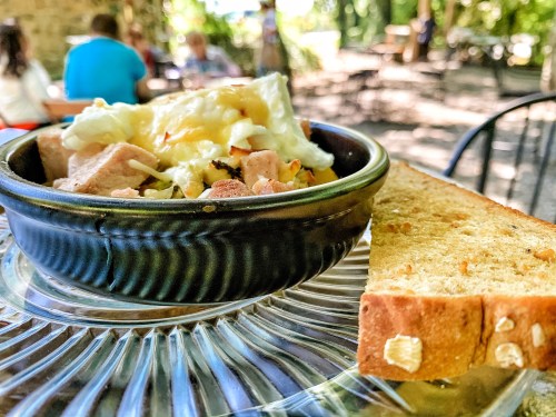 A bowl of hearty food with meat and vegetables, served with a slice of toasted bread, set in a sunny outdoor café.