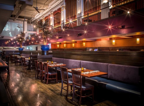 A stylish restaurant interior with wooden tables, chairs, and ambient lighting, featuring plants and a cozy atmosphere.