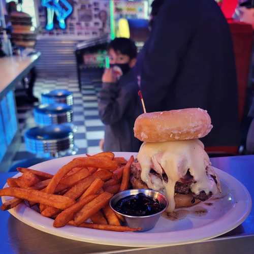 A burger topped with melted cheese and a donut, served with sweet potato fries and a small cup of sauce.