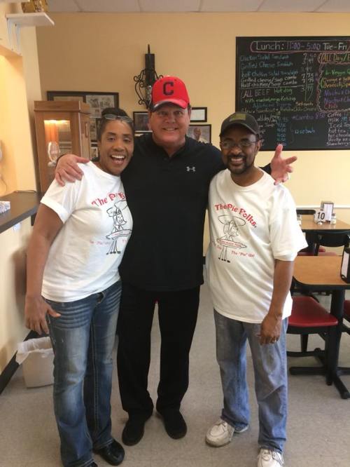 Three people smiling together in a casual restaurant setting, wearing matching t-shirts and a red baseball cap.