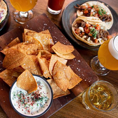 A wooden platter with crispy tortilla chips, a bowl of dip, tacos, and drinks on a table.