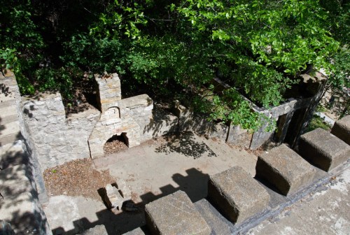 Ruins of a stone structure surrounded by greenery, with remnants of walls and a fireplace visible.