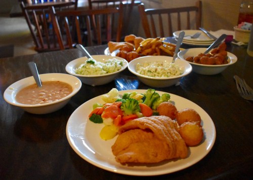 A plate of fried fish with vegetables and hushpuppies, surrounded by bowls of coleslaw, rice, and fried snacks.