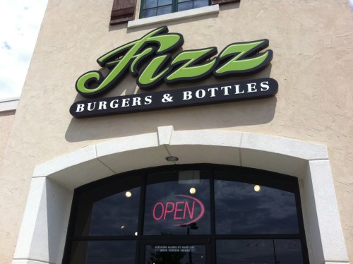 Sign for "Fizz Burgers & Bottles" with an open sign in the window, set against a light-colored building.