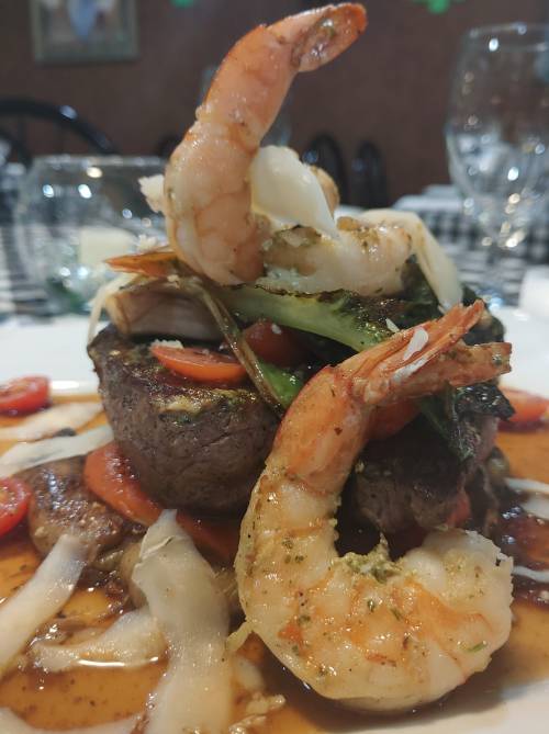 A gourmet dish featuring a steak topped with shrimp, vegetables, and a flavorful sauce, served on a white plate.