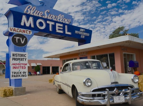 A vintage white car parked in front of the Blue Swallow Motel, featuring a retro sign and a clear blue sky.