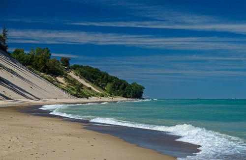 A serene beach scene with gentle waves, sandy shore, and lush green dunes under a clear blue sky.