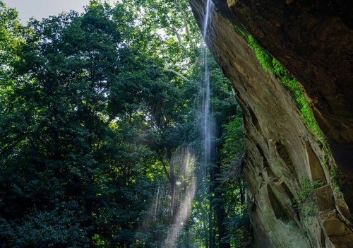 A serene waterfall cascading down a rocky cliff, surrounded by lush green trees and sunlight filtering through the foliage.