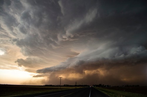 Dramatic storm clouds loom over a rural road, with dark skies and a hint of light on the horizon.