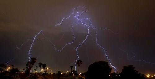 Dramatic lightning strikes illuminate a dark sky over a cityscape with palm trees and streetlights.