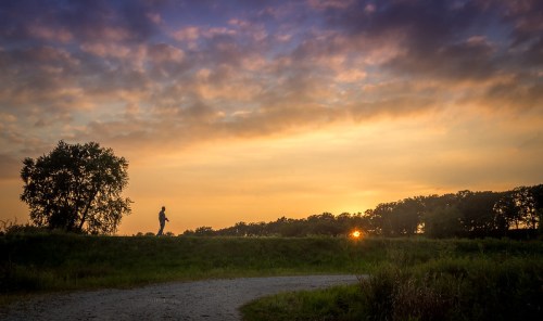 A person walks along a path at sunset, silhouetted against a colorful sky and trees in the background.