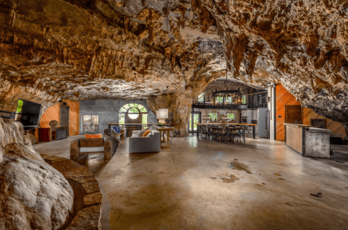 A spacious cave interior featuring modern furniture, a dining area, and a kitchen, with natural rock formations visible.