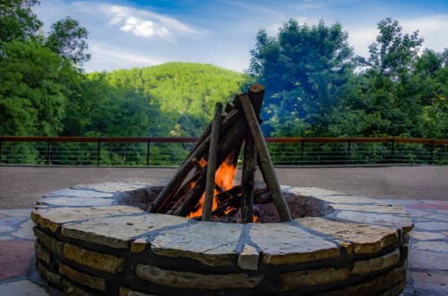A stone fire pit with a crackling fire, surrounded by lush green trees and a scenic mountain backdrop.