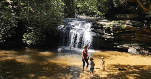 A serene waterfall cascades into a shallow pool, with children playing and adults enjoying the natural setting.