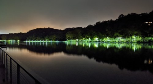 A calm lake at night, reflecting lights from the shore and surrounded by trees under a cloudy sky.