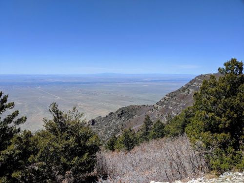 A panoramic view of a valley from a mountain, featuring clear blue skies and scattered trees on the slopes.