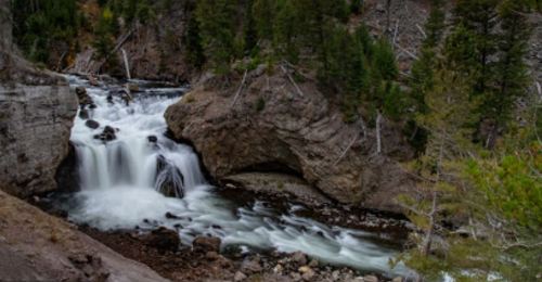 A serene waterfall cascades over rocky terrain, surrounded by lush green trees and rugged cliffs.