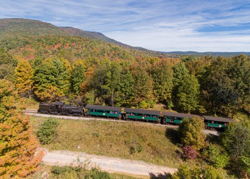 A vintage steam train travels through colorful autumn foliage in a scenic landscape.