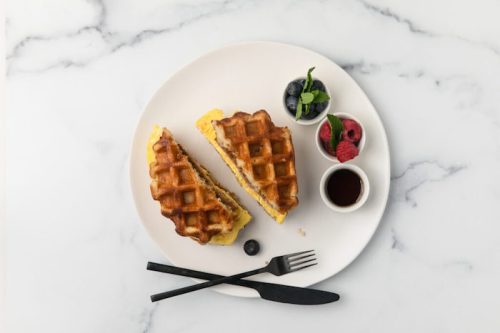 A plate with a waffle sandwich, fresh berries, and a small cup of syrup, set on a marble surface.
