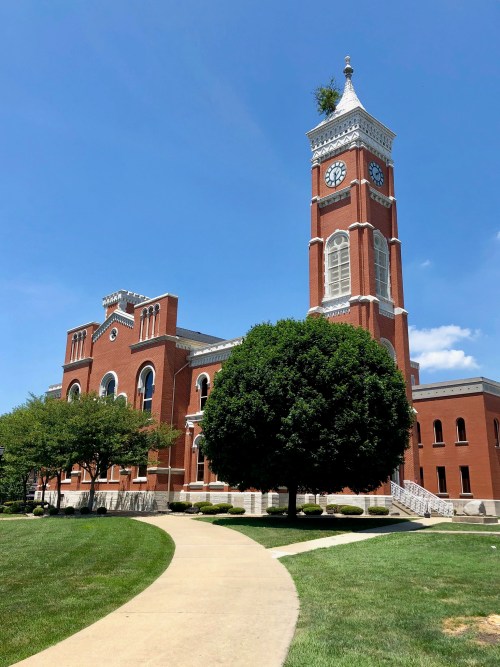 Historic brick building with a tall clock tower, surrounded by green grass and trees under a clear blue sky.