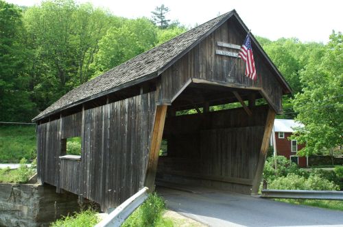 A rustic wooden covered bridge with an American flag, surrounded by lush green trees and a rural landscape.
