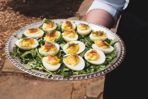 A silver platter holds deviled eggs topped with smoked salmon and dill, resting on a bed of arugula.
