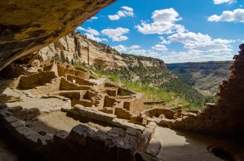 Ancient ruins nestled in a canyon, with a view of lush greenery and a blue sky dotted with clouds.