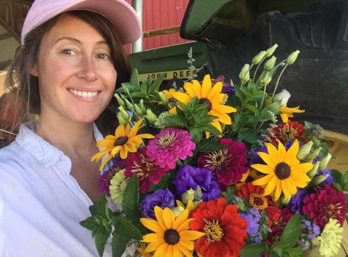 A woman in a pink cap smiles while holding a vibrant bouquet of colorful flowers.