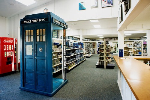 A blue police box stands in a store aisle filled with shelves of various products, with a soda machine in the background.