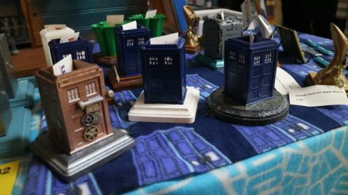 A display of miniature TARDIS models in various colors on a blue tablecloth, with other decorative items in the background.
