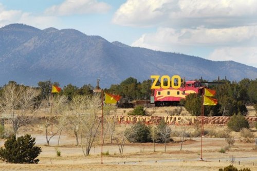 A colorful zoo sign stands against a backdrop of mountains and blue skies, with flags waving in the foreground.