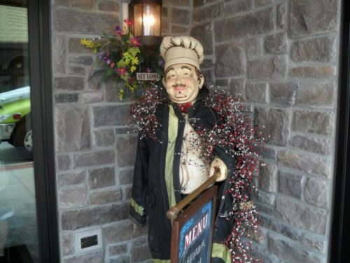 A decorative chef statue wearing a black coat, adorned with festive garlands, stands by a stone wall outside a restaurant.