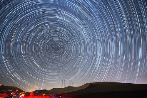 A starry night sky with circular star trails above dark hills and faint lights from vehicles in the foreground.