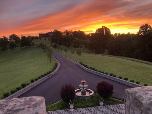 A scenic view of a winding driveway leading to a sunset over lush green fields and trees, with a fountain in the foreground.