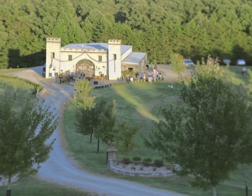 A scenic view of a castle-like building surrounded by trees, with people gathered outside on a grassy area.