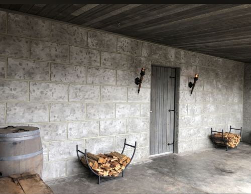 A stone wall with a wooden door, two wall sconces, and two wood holders filled with logs beside a barrel.