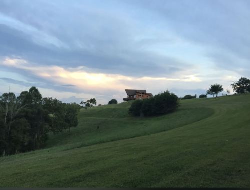 A serene landscape featuring a grassy hill with a house and trees under a cloudy sky at dusk.