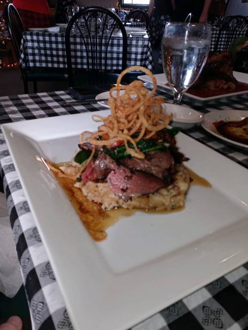 A plated dish featuring sliced steak on a bed of creamy risotto, topped with crispy onion rings and garnished with greens.