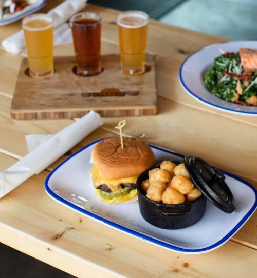 A cheeseburger with a pickle and a side of tater tots in a small cast-iron dish, with three beers in the background.