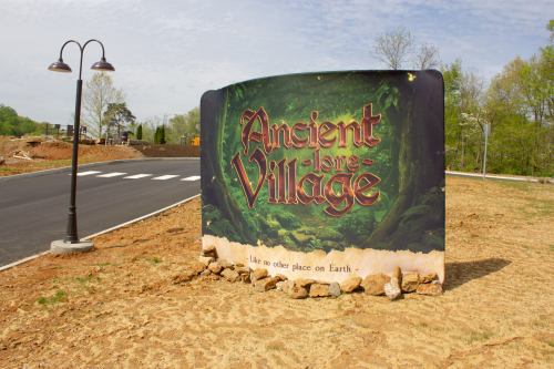 Sign for "Ancient Lore Village" with a tagline, set against a backdrop of trees and a newly paved road.
