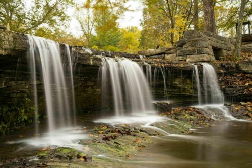 A serene waterfall cascades over rocky ledges, surrounded by autumn foliage and a tranquil pool below.